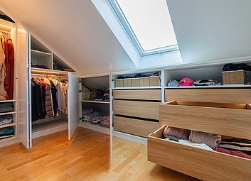 Begehbarer Kleiderschrank unter der Dachschräge mit geöffneten Türen und Schubkästen.