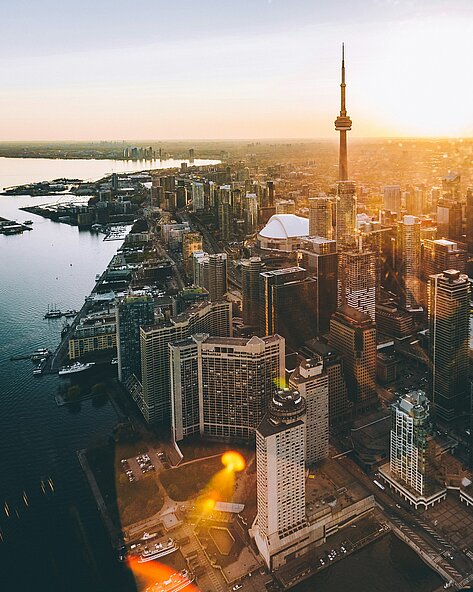 Luftaufnahme von Toronto mit CN Tower im Sonnenaufgang, Hafenanlagen und Hochhäusern entlang der Uferlinie.