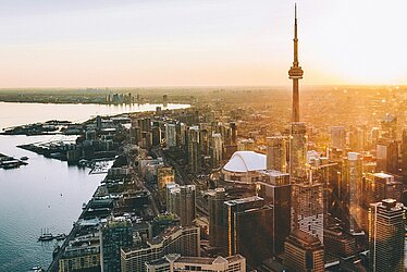 Luftaufnahme von Toronto mit CN Tower im Sonnenaufgang, Hafenanlagen und Hochhäusern entlang der Uferlinie.