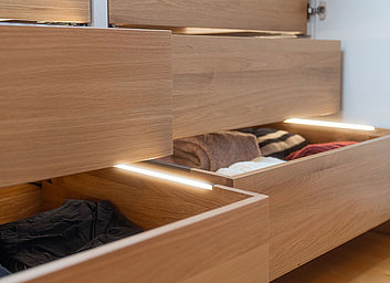 Begehbarer Kleiderschrank unter der Dachschräge mit geöffnetem Schubkasten und Beleuchtung im Schubkasten.