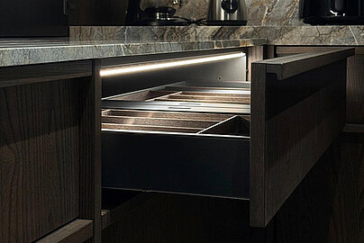 Geöffnete Küchenschublade mit beleuchtetem Innenraum und strukturierten Organisationselementen aus Holz und Metall